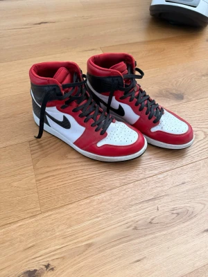 Nike Air Jordan 1 Retro High “Satin Snake Chicago” - Säljer ett par Nike Air Jordan 1 Retro High sneakers i klassisk röd, vit och svart färg. Skorna har högt skaft, svart swoosh och svarta skosnören. Ovandelen är i läder med perforerad tåbox för extra ventilation. Perfekta för dig som gillar streetwear och ikonisk sneakerstil.