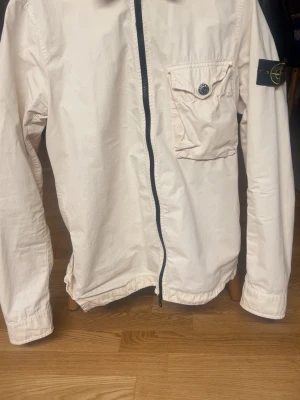 Beige overshirt från Stone Island - Snygg beige overshirt från Stone Island med svart dragkedja, bröstficka med knapp och klassisk logopatch på ärmen. Tillverkad i lätt bomullsmaterial som ger en clean och modern look. Perfekt att bära över en t-shirt för en avslappnad stil.