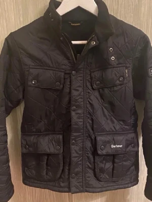 Svart quiltad jacka från Barbour - Stilren svart quiltad jacka från Barbour med hög krage, fyra fickor framtill och snygga detaljer som tryckknappar och Barbour-logga. Jackan har ett klassiskt mönster och justerbara spännen bak. Perfekt för dig som gillar tidlös design och funktion.