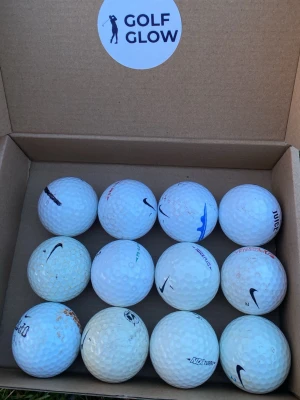 Golfbollar Nike och blandade märken - Säljer ett set med 12 vita golfbollar från Nike och andra märken, levereras i en kartong med Golf Glow-logga. Bollarna har klassisk rund form och dimpled yta, vissa med tryck och loggor i svart, blått och grönt. Perfekt för golfentusiasten som vill fylla på förrådet.