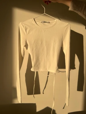 Ljus ribbad långärmad topp med snörning - Ljus långärmad topp med ribbad struktur och snörning i midjan. Toppen har en öppen rygg med snygga band som kan knytas och croppad passform. Perfekt för dig som gillar trendiga detaljer och stilren design.