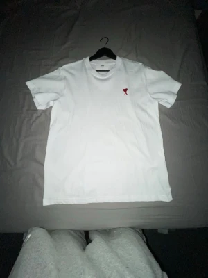 Vit t-shirt från Ami med hjärta - Vit t-shirt från Ami med klassisk passform och korta ärmar. På bröstet finns ett broderat rött hjärta med bokstaven A. Perfekt för dig som gillar stilrena plagg med en liten detalj. Materialet är mjuk bomull.
