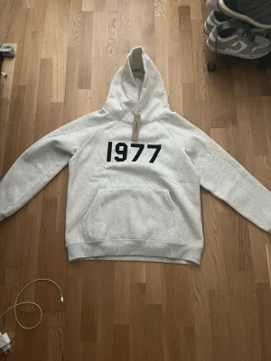 Grå hoodie med 1977-tryck - Snygg grå hoodie från Essentials med svart 1977-tryck på bröstet. Hoodien har en stor huva, ribbade muddar och en praktisk magficka. Perfekt för dig som gillar streetwear och enkel design. Materialet är mjukt och bekvämt, troligen bomullsblandning.