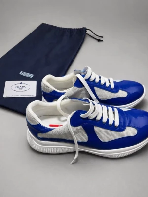 Blå och vita sneakers från Prada - Snygga sneakers från Prada med blå lackdetaljer och vita meshpaneler. Skorna har vita snören och en vit sula med röda detaljer undertill. Perfekt för dig som vill sticka ut med ett exklusivt märke och cool design.