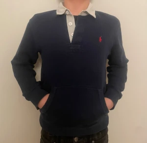 Polo Ralph Lauren tröja  - Mörkblå långärmad tröja från Ralph Lauren med vit krage och knappslå. Tröjan har en röd broderad logga på bröstet och en stor magficka. Perfekt för en avslappnad och stilren look. Det är ett diskret hål i ena ärmen som knappt syns se bild nr 