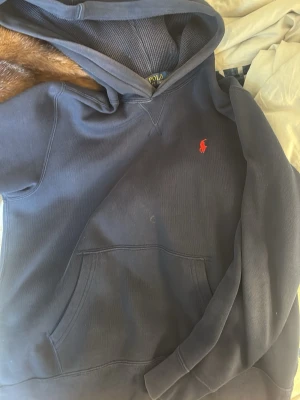 Mörkblå hoodie från Polo Ralph Lauren - Mörkblå hoodie från Polo Ralph Lauren med klassisk röd broderad logga på bröstet. Hoodien har stor känguruficka framtill och ribbade muddar vid ärmar och nederkant. Tillverkad i mjukt bomullsmaterial och har en skön huva.
