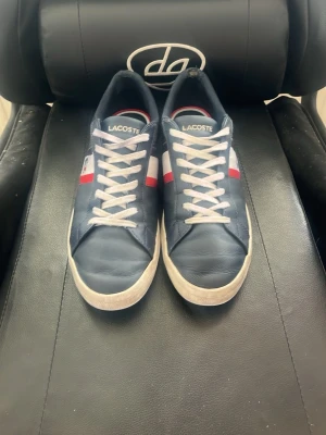 Blå Lacoste sneakers med vita detaljer - Snygga blå sneakers från Lacoste med vita och röda detaljer på sidan samt vita skosnören. Klassisk låg modell med Lacoste-logga på sidan och tungan. Perfekt för dig som vill ha en stilren och sportig look.