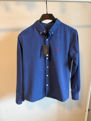 Blå skjorta från Polo Ralph Lauren - Klassisk blå skjorta från Polo Ralph Lauren med röd broderad logga på bröstet. Skjortan har lång ärm, knappar framtill och button-down krage. Tillverkad i mjuk bomull och har en normal passform. Perfekt för en stilren och tidlös look.