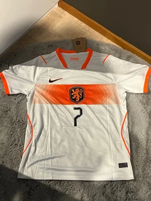 Vit Nederländerna fotbollströja Nike - Snygg vit fotbollströja från Nike med orange detaljer och Nederländernas landslagsemblem på bröstet. Nummer 7 och namnet Xavi på ryggen. Tröjan har korta ärmar, V-ringning och ventilerande material, perfekt för träning eller match.