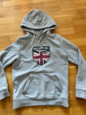 Grå Morris hoodie XS - Snygg grå hoodie från Morris med stor Union Jack-flagga och texten 'Morris Est. 1968' på bröstet. Klassisk känguruficka framtill, justerbar dragsko i huvan och mjukt material. Perfekt för en avslappnad streetwear-look. Skicket är fint, mått står på bilderna. Priset kan förändras vid snabb affär✅👍