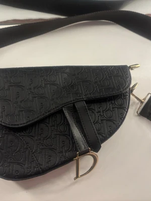 Svart Saddle Bag i läder - Säljer en svart Saddle Bag Väskan har guldfärgade metalldetaljer och ett stort D-hänge framtill. Snygg axelrem och unik form som verkligen sticker ut.