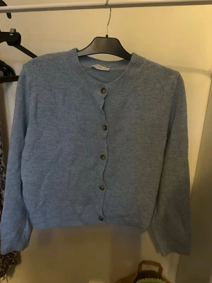 Ljusblå stickad kofta med knappar - Mysig ljusblå stickad kofta med rund halsringning och metallfärgade knappar framtill. Koftan har långa ärmar och ribbade muddar vid ärmslut och nederkant. Perfekt att slänga på sig över en t-shirt eller klänning.