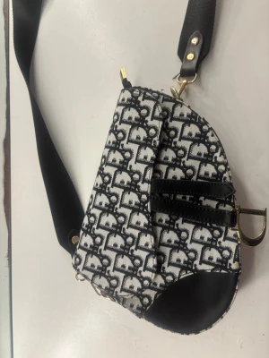 Svartvit axelväska med monogram - Snygg axelväska i svartvitt tyg med monogrammönster och detaljer i svart läder. Väskan har guldfärgade metalldetaljer, bred axelrem och unik form. Perfekt för dig som vill ha en trendig accessoar med statement-känsla.