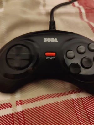 Sega Mega Drive handkontroll svart - Klassisk svart handkontroll till Sega Mega Drive med rundad form, D-pad, tre knappar och en röd startknapp. Perfekt för retrospel och nostalgiska gaming-sessioner. Kontrollen är tillverkad i plast och har kabelanslutning.