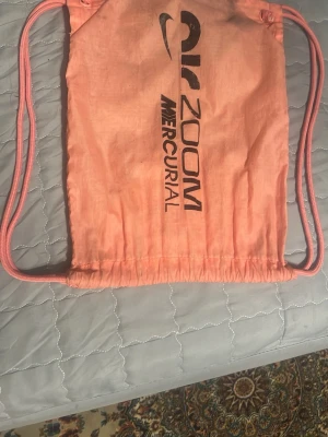 Nike Zoom Mercurial gympåse - Cool gympåse från Nike i en ljusrosa färg med svarta tryck på båda sidor. Påsen har snörstängning och är tillverkad i lätt syntetmaterial. Perfekt för att bära träningskläder eller skor. Designen är enkel och sportig med Nike-logga och Mercurial-tryck.