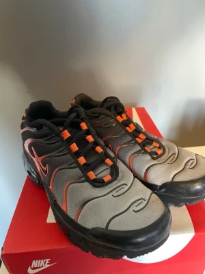Nike Air Max Plus svart och orange - Nike Air Max Plus sneakers i svart och orange med tydliga TN-loggor på sulan. Skorna har en unik vågig design på hälen och meshmaterial upptill. Sulan har synliga Air Max-bubblor och mönster i rött och svart. Perfekt för dig som gillar sportiga och coola sneakers.