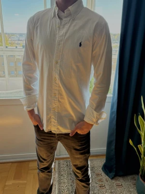 Vit polo Ralph louren skjorta - Färsch vit skjorta från Ralph Lauren med den ikoniska blå logotypen broderad på bröstet. Skjortan har lång ärm, knappar framtill och är tillverkad i mjuk bomull. Perfekt för en clean och stilren look nu till sommaren!                                         PRIS KAN DISKUTERAS VID SNABB AFFÄR!
