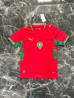 Marockansk landslagströja Puma - Säljer en röd Marockansk landslagströja från Puma med gröna detaljer på ärmar och sidor. Tröjan har landslagets emblem på bröstet och gul Puma-logga. Materialet är lätt och andas, perfekt för fotboll eller träning.