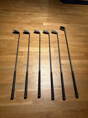 Set med 6 golfklubbor i metall - Säljer ett set med sex golfklubbor i metall med svarta grepp. Klubborna har klassisk form och passar för dig som vill komplettera din golfutrustning. Perfekt för både träning och spel på banan. Inget märke synligt på klubborna.