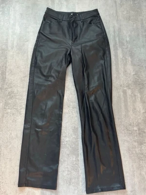 Svarta raka jeans i skinnlook - Säljer ett par svarta jeans med skinnlook och raka ben. De har klassisk femficksdesign, hög midja och stängs med knapp och dragkedja. Perfekta för dig som vill ha en edgy och trendig stil. Är sönder i en ögla där man trär in skärpet men inget man tänker så mycket på.