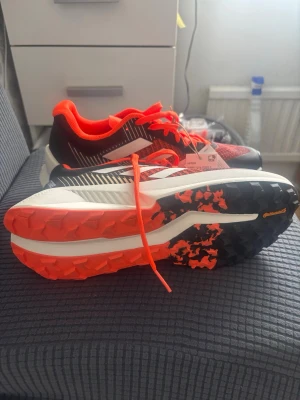 Adidas röda och svarta sneakers - Snygga sneakers från Adidas med röd och svart mesh-överdel, vita detaljer och orangea skosnören. Yttersulan har en cool mix av svart och orange mönster med Continental-gummi för extra grepp. Perfekt för dig som gillar sportig stil och komfort.