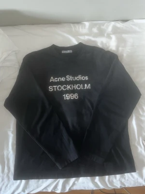 Svart långärmad tröja Acne Studios - Svart långärmad tröja från Acne Studios med vit text 'Acne Studios STOCKHOLM 1996' på bröstet. Tröjan har rund hals och är tillverkad i mjukt material, perfekt för en cool och avslappnad look.