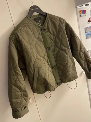 Olivgrön quiltad vindjacka från Zara - Säljer en olivgrön quiltad vindjacka från Zara med rund halsringning och stora knappar framtill. Jackan har ett unikt mönster, två fickor och dragsko i nederkant. Perfekt för dig som gillar en avslappnad och trendig look.