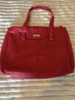 Röd väska  - Stilren röd väska med dubbla axelband och en metallbricka framtill. Väskan har enkel design med dragkedja upptill och gott om plats för allt du behöver. Perfekt för dig som vill ha en snygg och praktisk väska.