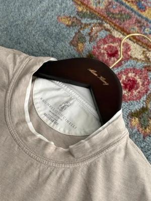 Brunello Cucinelli T-shirt - Stilren beige t-shirt från Brunello Cucinelli med klassisk rund halsringning och regular fit. Tillverkad i mjuk bomull som känns skön mot huden. Perfekt basplagg för en clean och avslappnad look. Nypris 4500kr, tvättlappen är bortsprättad men annars som ny