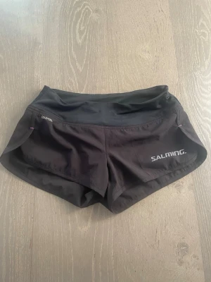 Svarta träningsshorts från Salming - Snygga svarta träningsshorts från Salming med logga på vänster ben och bred låg midja för extra komfort. Tillverkade i ett lätt och snabbtorkande syntetmaterial, perfekta för intensiva träningspass. Sportig och tight passform.