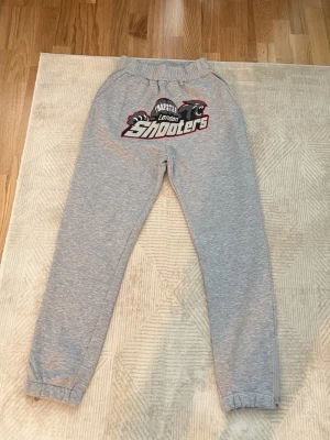 Gråa Trapstar London Shooters mjukisbyxor - Säljer ett par gråa mjukisbyxor från Trapstar med stort broderat London Shooters-motiv framtill. Byxorna har resår i midjan och vid bensluten samt sidofickor. Perfekta för en chill och sportig look.