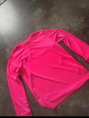 Rosa långärmad Nike topp - Säljer en snygg långärmad topp från Nike i stark rosa färg. Tröjan har rund halsringning och är tillverkad i ett mjukt, stretchigt syntetmaterial som passar perfekt till träning eller chill. Liten Nike-logga på bröstet för en sportig vibe.