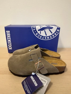 Birkenstock Boston taupe mockatofflor - Säljer ett par Birkenstock Boston tofflor i taupefärgad mocka med justerbart spänne i metall. Klassisk slip-in modell med mjuk fotbädd och korksula för extra komfort. Perfekta för dig som vill ha både stil och bekvämlighet.