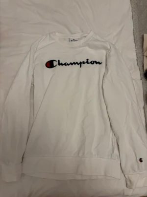 Vit sweatshirt från Champion - Vit sweatshirt från Champion med klassiskt broderat logotyptryck på bröstet och liten logga på ärmen. Tröjan har rund halsringning, långa ärmar och ribbade muddar. Perfekt för en avslappnad och sportig look. Tröjan har en gräsfläck på armbågen och en liten fläck längst ner på ryggen, som ni kan se på bilderna. Tröjan är i barnstorlek för 11/12 år och 150/155 cm.