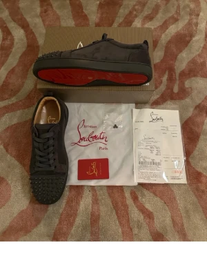 Christian Louboutin gråa sneakers - Gråa sneakers från Christian Louboutin med klassiska röda sulor och nitar på tån. Skorna har snörning och är tillverkade i mocka med läderdetaljer. Perfekt för dig som vill sticka ut med exklusiva och edgy skor.