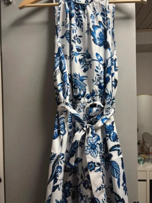 Blåvit blommig klänning med knyt - Snygg blåvit klänning med blommigt mönster och knytband i midjan. Klänningen är ärmlös och har en luftig passform som passar perfekt till varma dagar. Materialet känns mjukt och behagligt mot huden.