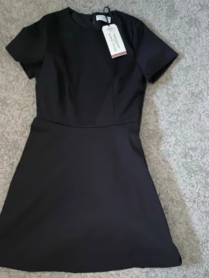 Svart kortklänning Holly & Whyte - Stilren svart kortklänning från Holly & Whyte med rund halsringning och korta ärmar. Klänningen har en lätt utställd kjol och är tillverkad i en återvunnen polyesterblandning. Perfekt för dig som gillar enkel och klassisk stil.