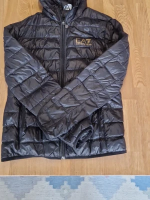 Svart pufferjacka från EA7 Emporio Armani - Säljer en svart pufferjacka från EA7 Emporio Armani med quiltad design och huva. Jackan har dragkedja framtill och logotyp på bröstet. Materialet är glansigt och lätt, perfekt för kalla dagar. Stilsäker och funktionell jacka som håller dig varm.