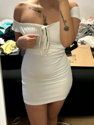 Vit off shoulder ribbad klänning - Trendig vit off shoulder klänning med ribbad struktur och knytning framtill. Kort, figurnära modell som framhäver siluetten. Materialet känns mjukt och stretchigt.