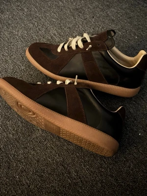 Maison Margiela sneakers brun/svart - Maison Margiela sneakers i brun och svart med beige snörning och gummisula. Skorna har en klassisk låg profil och kombinerar läder och mocka för en stilren look. Perfekta för dig som vill ha något unikt och trendigt.