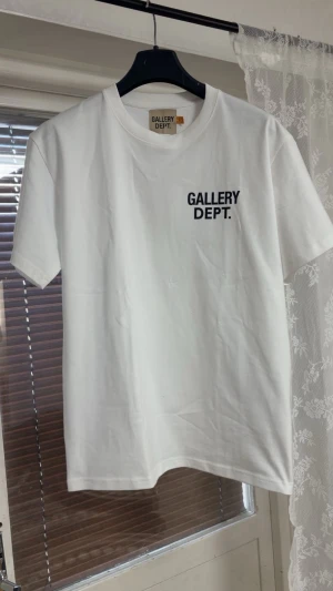 Vit t-shirt från Gallery Dept. - Vit t-shirt från Gallery Dept. med svart tryck på bröstet och stort tryck på ryggen med texten 'GALLERY DEPT. HOLLYWOOD CA.'. Klassisk rund hals och korta ärmar. Perfekt för dig som gillar streetwear och enkel stil.