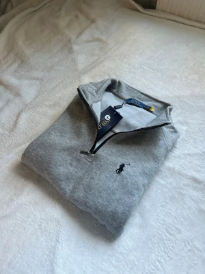 Grå quarter zip tröja från Polo Ralph Lauren - Snygg grå tröja med quarter zip från Polo Ralph Lauren. Tröjan har en klassisk krage och det ikoniska broderade logotypen på bröstet. Perfekt för lager-på-lager och tillverkad i mjukt material som känns skönt mot huden.