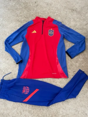 Spaniens träningsset Adidas röd/blå - Säljer ett fett träningsset från Adidas med Spaniens landslagslogga. Tröjan är röd med blå ärmar och har halv dragkedja, medan byxorna är blå med röd detalj och logga. Perfekt för fotbollsträning eller chill. Materialet är funktionspolyester som andas.