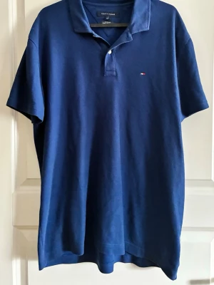 Marinblå pikétröja Tommy Hilfiger - Klassisk marinblå pikétröja från Tommy Hilfiger med liten logga på bröstet. Tröjan har korta ärmar, krage och knappar vid halsen. Perfekt till jeans eller shorts för en avslappnad men snygg look. L men sitter mer som M