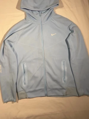 Nike NOCTA ljusblå  - Ljusblå mjukisbyxor från Nike NOCTA med raka ben och dragsko i midjan. Diskreta vita loggor och textdetaljer på benen. Perfekt för chill och streetwear. Materialet är mjukt och bekvämt, troligen bomull eller polyester.