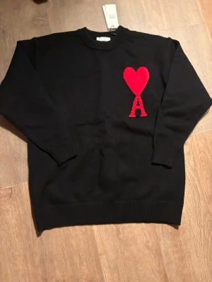 Svart stickad tröja från Ami - Svart stickad tröja från Ami Alexandre Mattiussi med röd hjärt- och A-detalj på bröstet. Tröjan har rund halsringning, ribbade muddar och nederkant. Perfekt för dig som gillar stilrena plagg med en twist.
