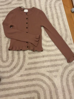 Brun ribbad kofta med volangkant - Snygg brun ribbad kofta med knappar framtill och volangkant vid nederdelen och ärmslut. Långärmad och figurnära modell, perfekt för att skapa en trendig look. Materialet är mjukt och stretchigt, vilket gör den bekväm att bära.