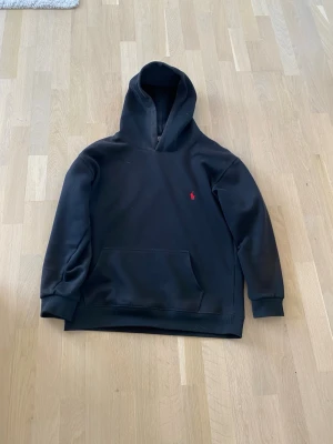 Svart hoodie från Polo Ralph Lauren - Det står att det är storlek XL i barn som är M i vuxen men den är som en S i vuxen. Svart hoodie från Polo Ralph Lauren med klassisk huva och stor magficka. Diskret röd logga broderad på bröstet. Mjukt material som känns skönt mot huden och ribbade muddar vid ärmslut och nederkant. Perfekt för en avslappnad och cool look. 