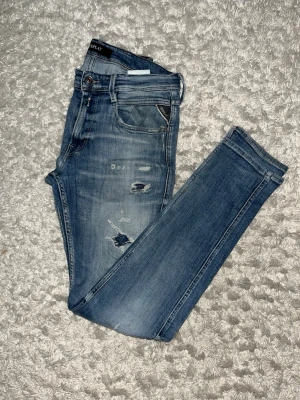 Blå slitna jeans från Replay - Säljer ett par blå jeans från Replay med slim fit och slitningar på både lår och knän. Jeansen har klassiska fem fickor, dragkedja och knappstängning. Materialet är denim med snygg tvätt och distressed detaljer. W32 L34