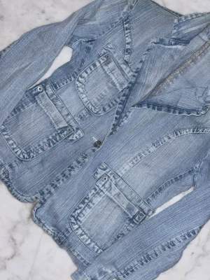 Ljusblå jeansjacka med fickor - Cool ljusblå jeansjacka med två stora fickor framtill och knappar både på ärmslut och fram. Jackan har en klassisk krage och snygga sömdetaljer som ger den en unik look. Perfekt för dig som gillar denim och vill ha något som sticker ut.